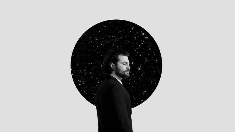 A man in a dark suit stands in profile against a minimalist gray background. Behind him, a circular black portal filled with stars evokes a cosmic, introspective atmosphere, suggesting themes of existentialism and inner depth.